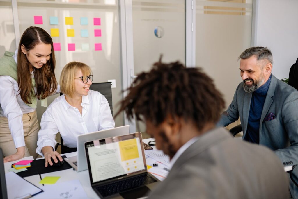 Vier collega’s in overleg tijdens een werkvergadering; twee vrouwen en twee mannen zitten en staan rondom een tafel met laptops, notities en gekleurde post-its op de achtergrond.